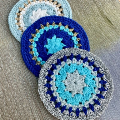 3 crochet coasters in shades of blue with silver and gold sparkle yarn