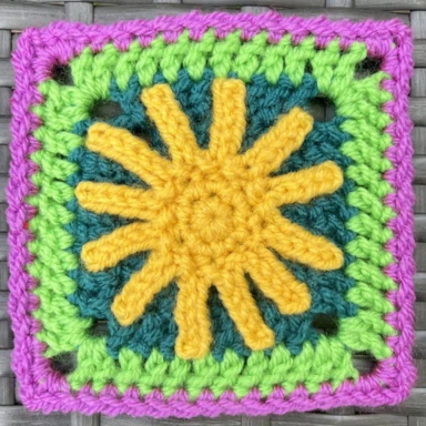 The sunny say crochet granny square with a bright yellow sun in the centre and surrounded by rows of teal, bright green and pink colours.