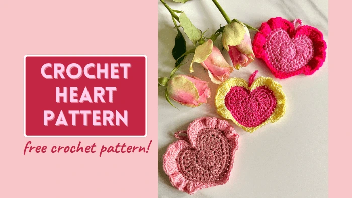 3 crochet love hearts in various shades of red and pink, placed on a table with 3 yellow and pink roses laid next to the hearts