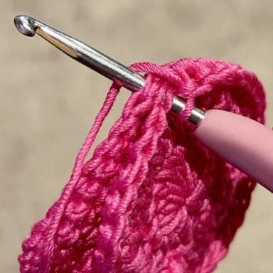 Pink crochet hook inserted into 2 adjacent crochet stitches using pink yarn