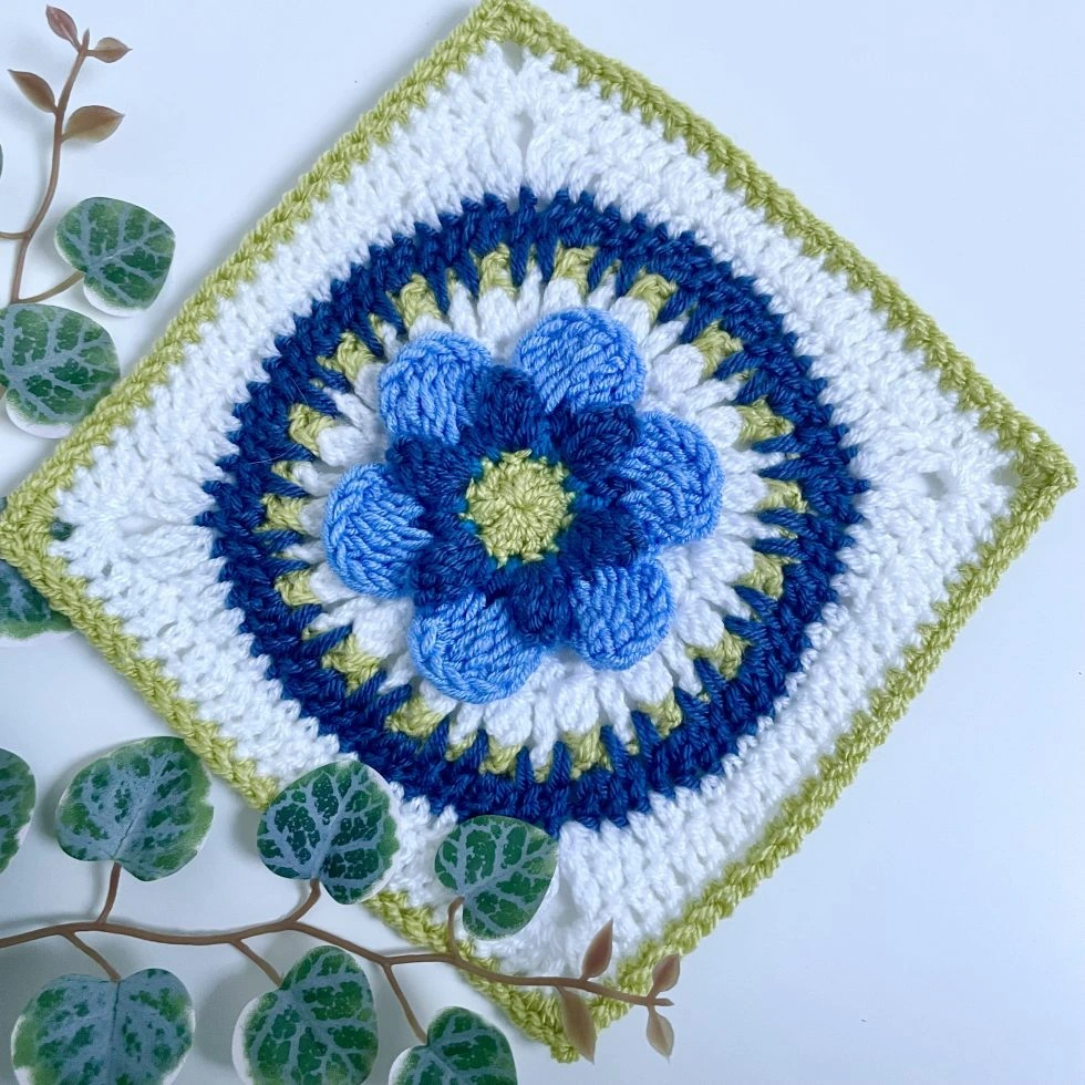 Crochet square with a flower in the centre, made with white, dark blue, lighter blue and a leaf green yarn. There are some leaves in the background