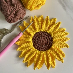 Crochet sunflower with a crochet hoot, scissors and 2 balls of yarn in yellow and brown which are the same colours used to make the sunflower